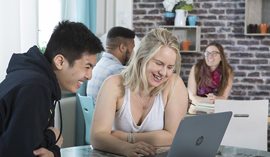 Kaksi opiskelijaa katsoo yhdessä hymyillen kannettavaa tietokonetta. Taustalla istuu muita opiskelijoita.