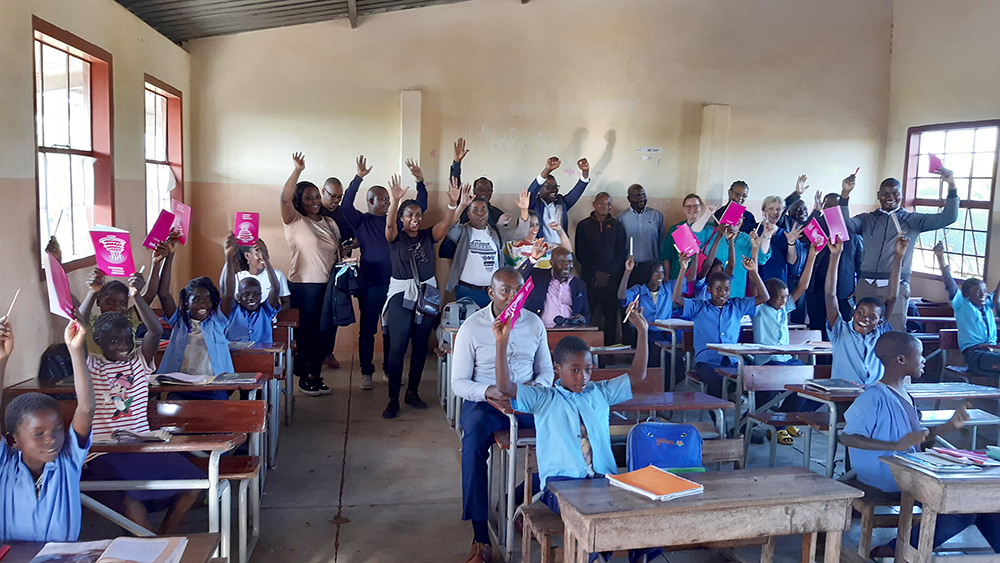 Mosambikilaisen kyläkoulun luokassa ja alakouluikäisiä koululaisia sekä luokan taustalla projektissa työskenteleviä henkilöitä nostaa kätensä ilmaan. Lapsilla käsissään vihkoja.