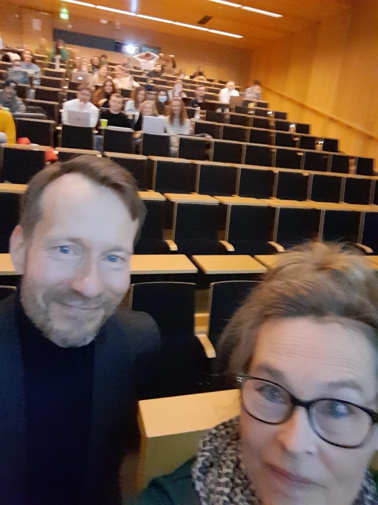 Asiantuntija Jyrki Ruohomäki ja lehtori Pia Viklund luentosalin etuosassa hymyilemässä kameralle. Heidän takanaan näkyy luennolle osallistuneita opiskelijoita luentosalin katsomossa. 